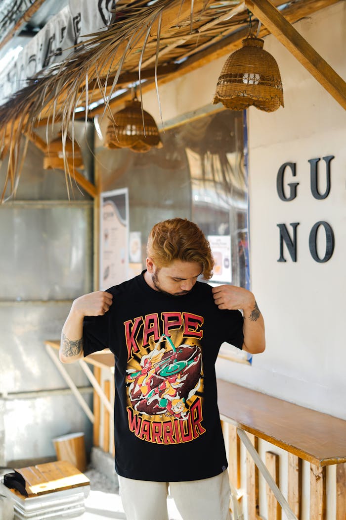 Home A young man wearing a graphic Kape Warrior t-shirt stands outdoors, checking his shirt design.