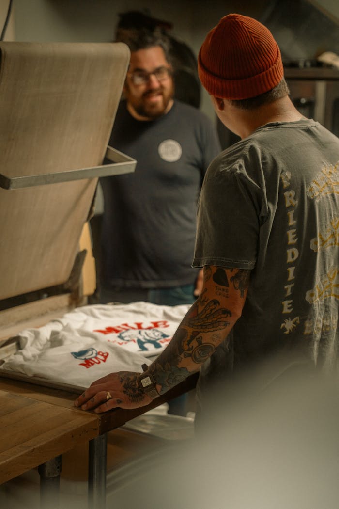 Home Two men engaged in screen printing a t-shirt in a cozy workshop setting.